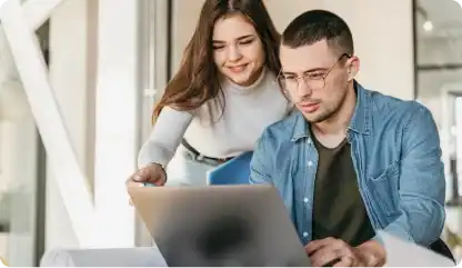 Mulher branca com cabelo cor castanho vestindo uma blusa de manga longa com gola alta junto com um homeme branco usando oculos com cabelo preto cortado vestindo blusa jeans de manga longa com camisa preta por baixo olhando para o computador 